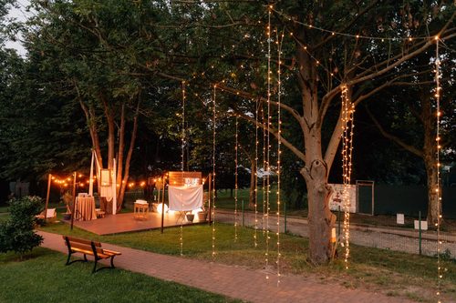 The park at dusk: trees strung with lights and a timber deck set for a ceremony